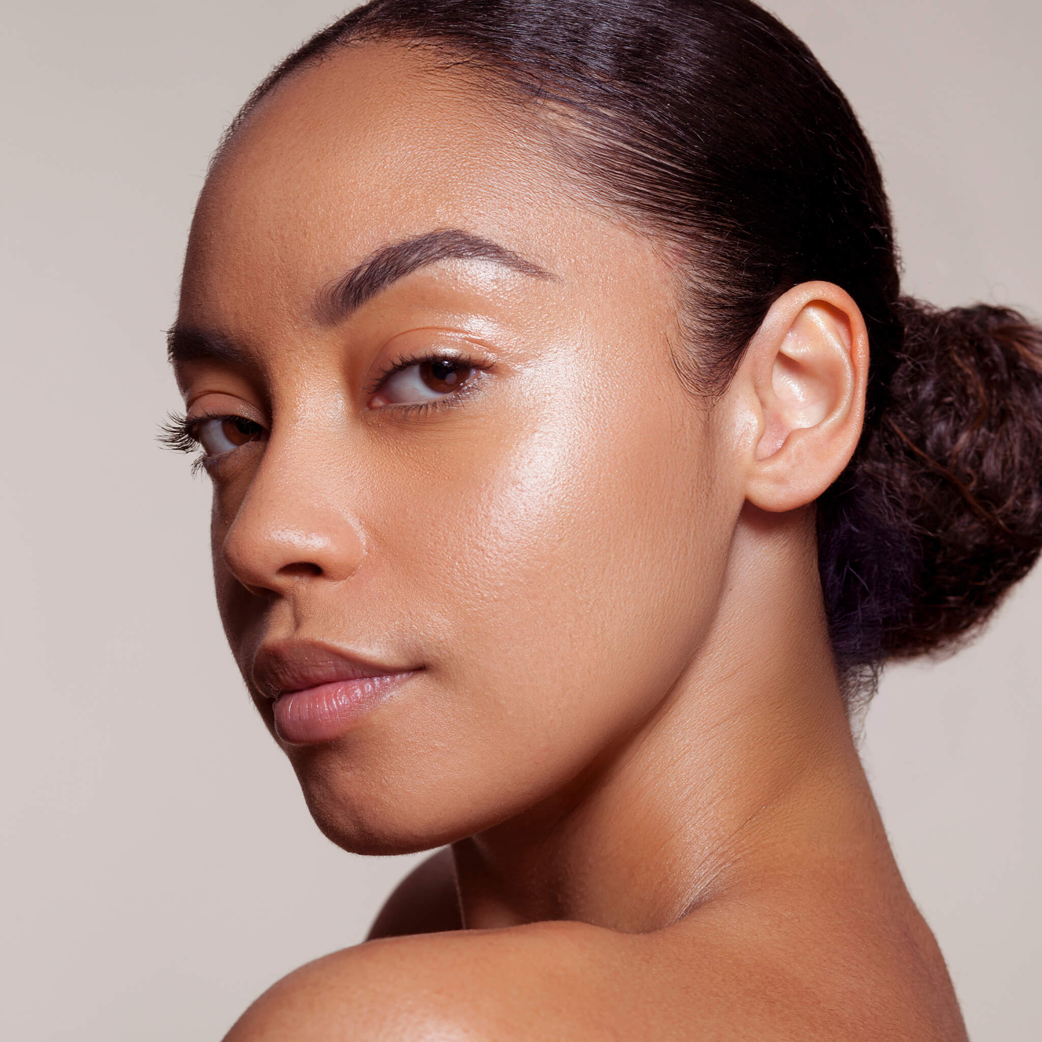Close-up portrait of a woman with luminous, healthy-looking skin and sleek pulled-back hair against a soft neutral background. Minimal makeup with a natural glow highlighting the complexion and facial contours.
