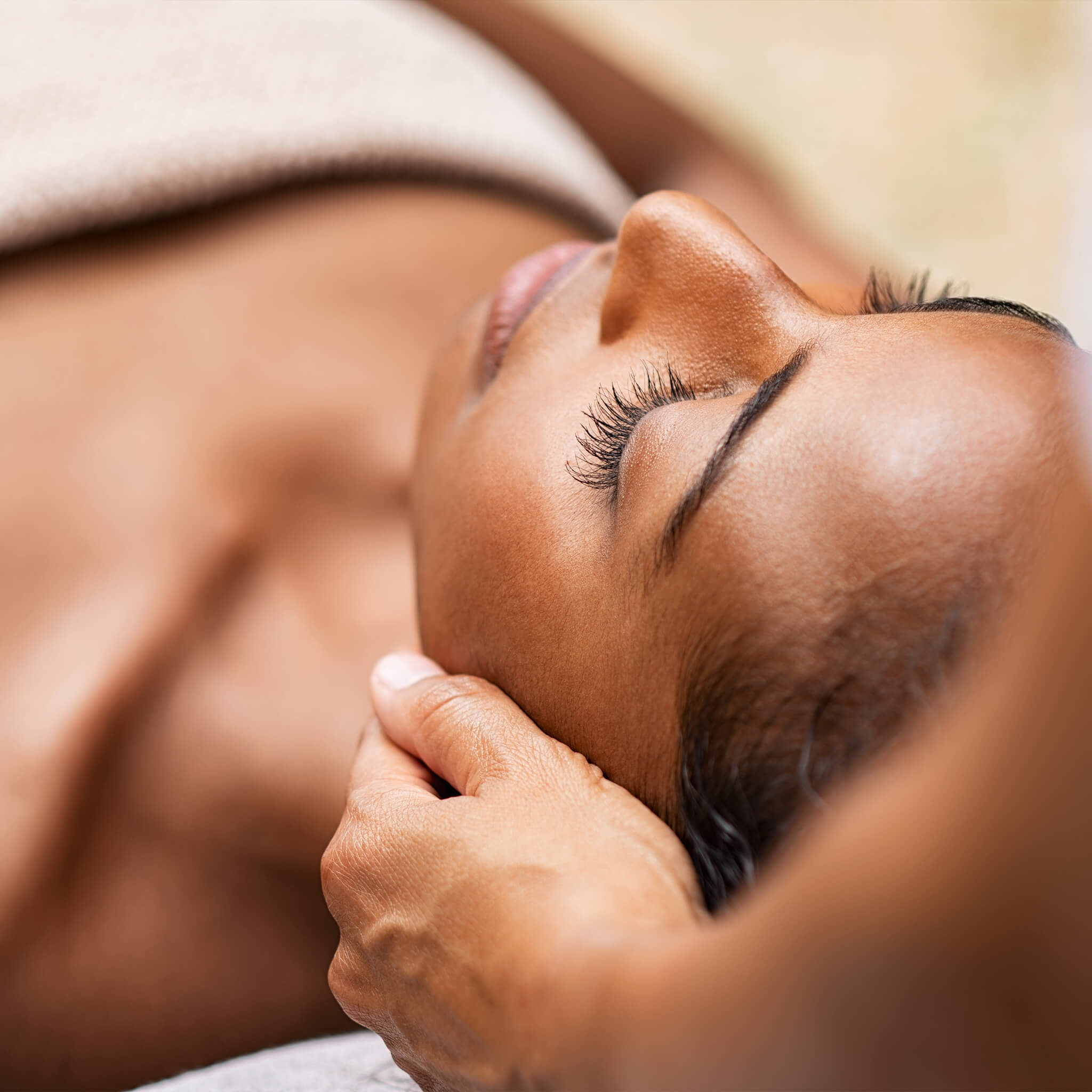 Close-up of a woman receiving a relaxing facial treatment with eyes closed, glowing skin and hands gently supporting the face in a calm aesthetic clinic setting.