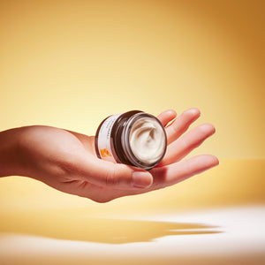 A hand holds an open jar of pale yellow face cream