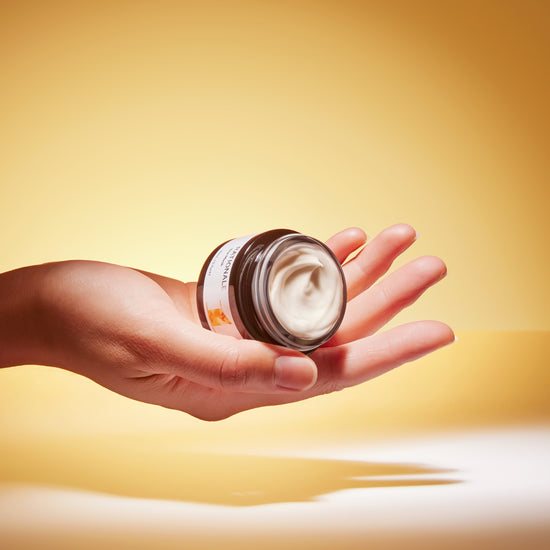 A hand holds an open jar of pale yellow face cream