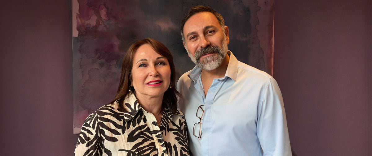 Dr Joseph Hkeik and Aesthetic Nurse Sally standing together in front of a dark purple wall with a painting behind them. The woman is wearing a black and white patterned top, and the man is wearing a light blue button-down shirt with glasses hanging from the collar.