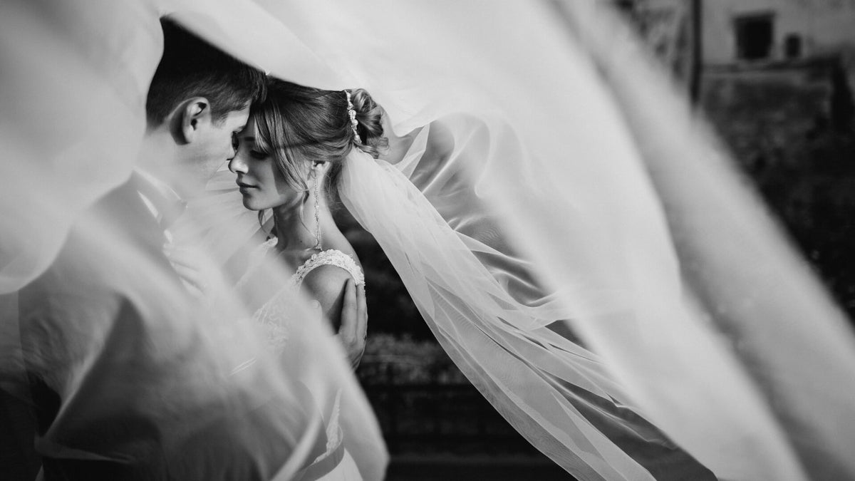 Bride and groom in soft focus, veil and lace dress draped as they hold hands in a calm outdoor setting