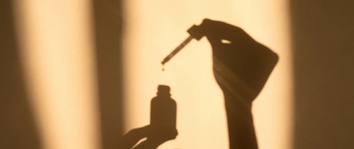A warm, backlit silhouette of a hand holding a dropper above a small skincare bottle, with a single drop hanging between the dropper and bottle.
