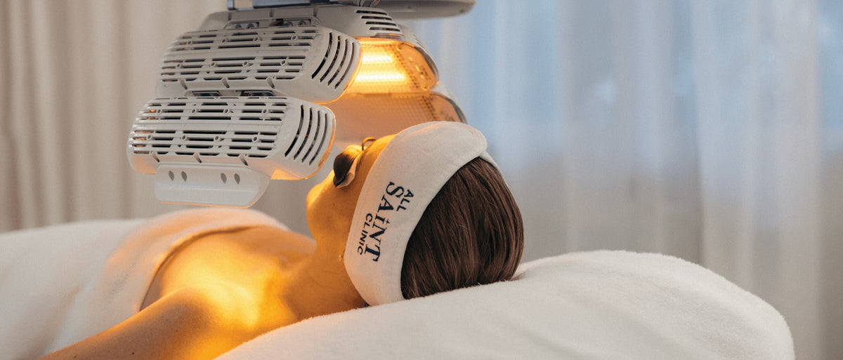A woman lying on a treatment bed under a glowing LED light machine, wearing a white headband with the brand name 'Saint' printed on it.