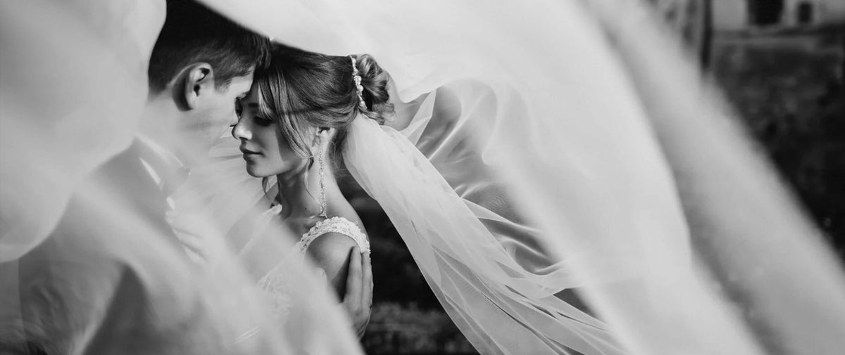 Black and white wedding photograph showing a bride with flawless skin in a sheer veil embracing a groom, with soft fabric folds framing the couple.