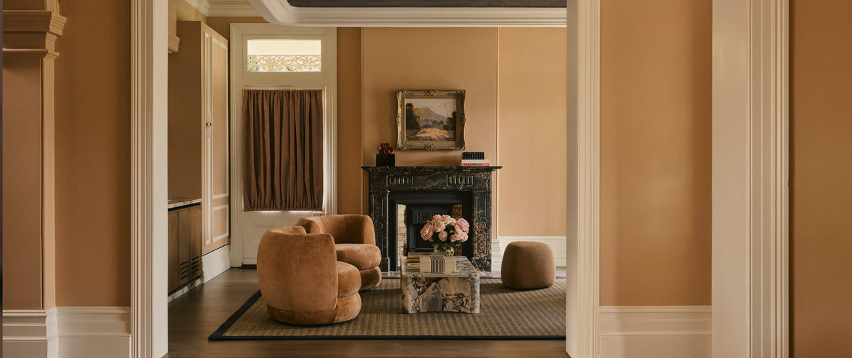 A styled interior room scene with warm tan walls, a dark fireplace, a round tan armchair, a small glass coffee table with flowers, and a textured rug, viewed through an open doorway.
